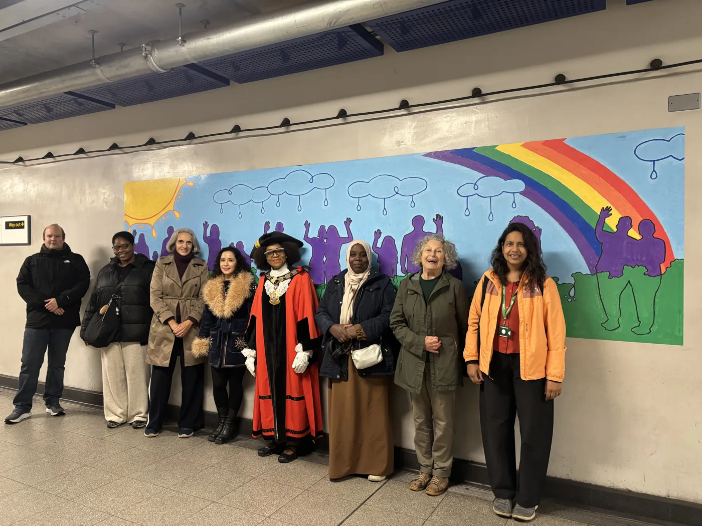 Eight people, including the Mayor of Islington in red robes and a tricorn hat, standing in a Tube station corridor in front of a mural of purple silhouettes waving under a sun, rain clouds and a rainbow