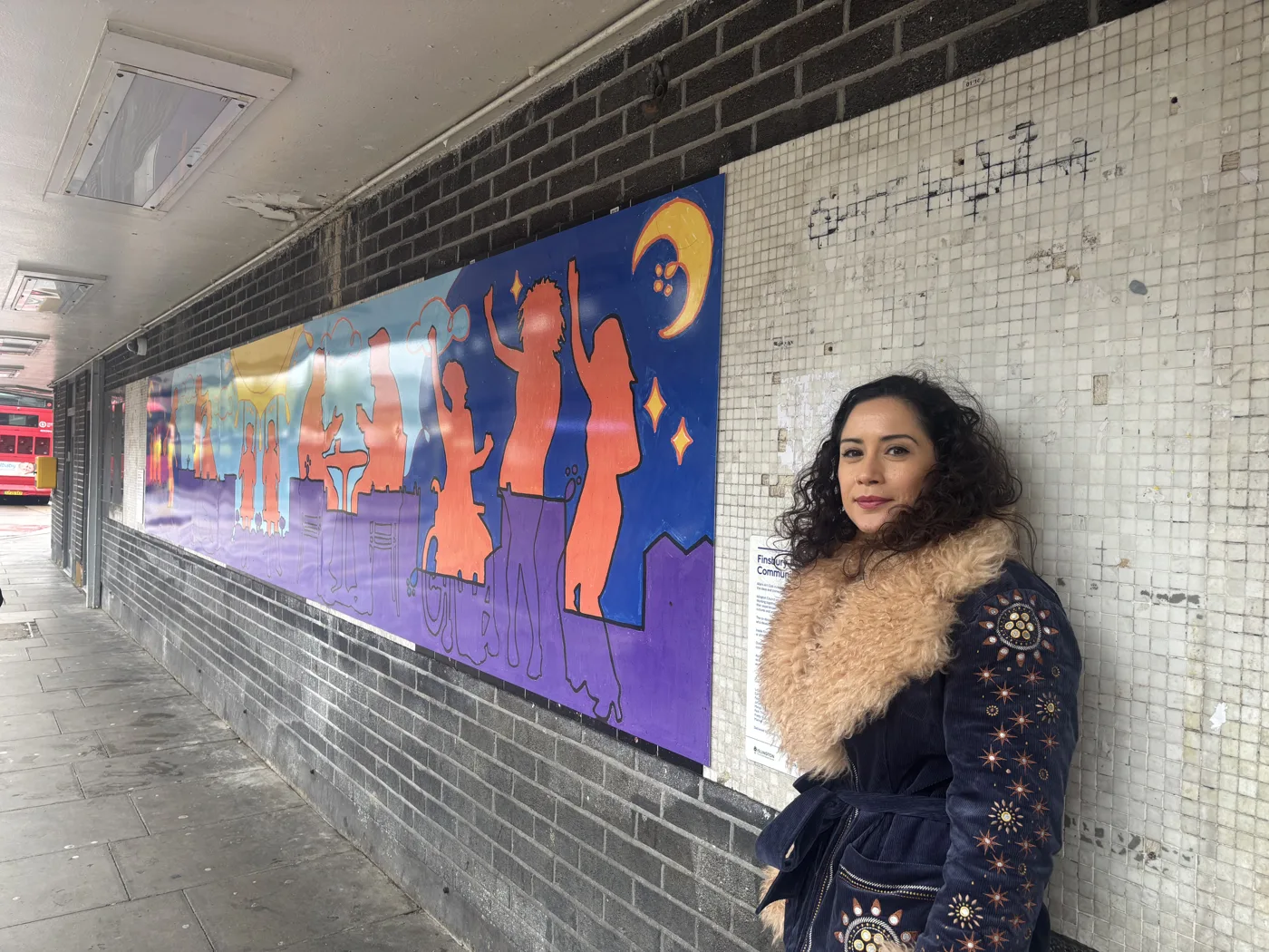 Lydia Rosen, in a navy coat with a fur collar, standing beside the Wells Terrace mural of orange silhouettes against a blue and purple night sky with a crescent moon, with a red bus in the distance