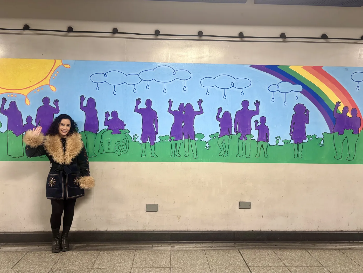 Lydia Rosen waving in front of the mural inside Finsbury Park station, a row of purple silhouettes of people with their hands raised on green grass under a sun, clouds and a rainbow