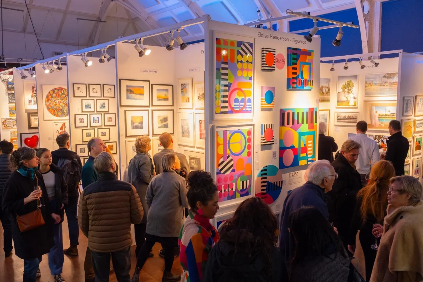 A crowd of visitors among white display walls hung with framed artworks in a hall with a glazed roof