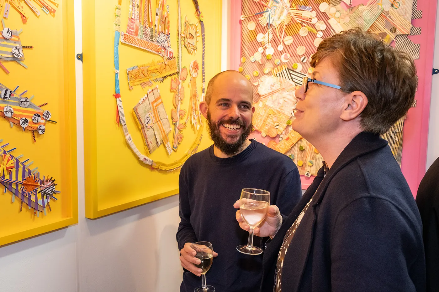 Two visitors with drinks talking in front of a wall of colourful work at a previous Highgate Art Fair