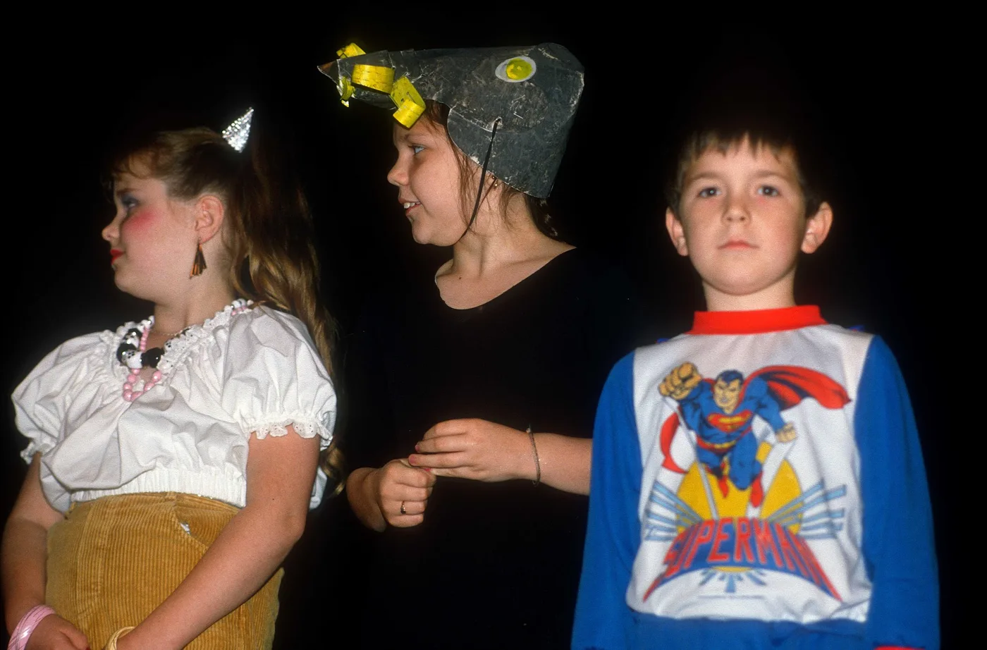 Three children standing against a dark background, one wearing a Superman top
