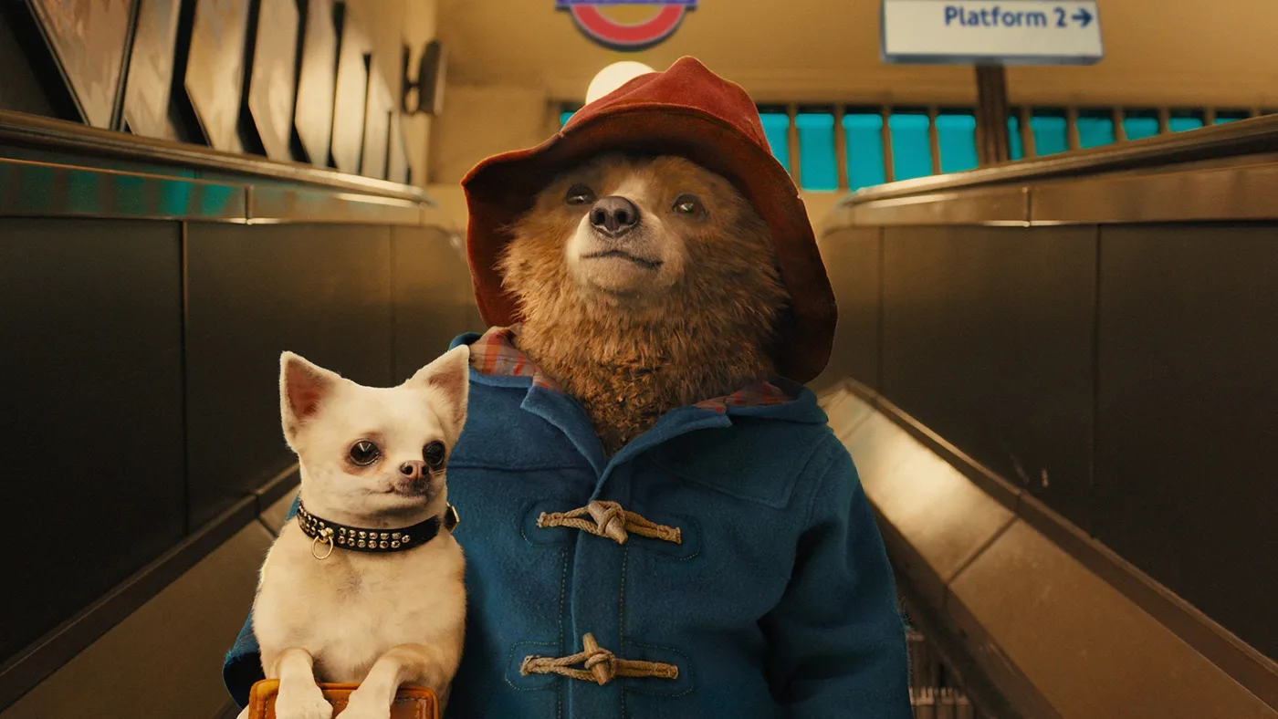 Paddington Bear in his red hat and blue duffel coat beside a small dog on an escalator