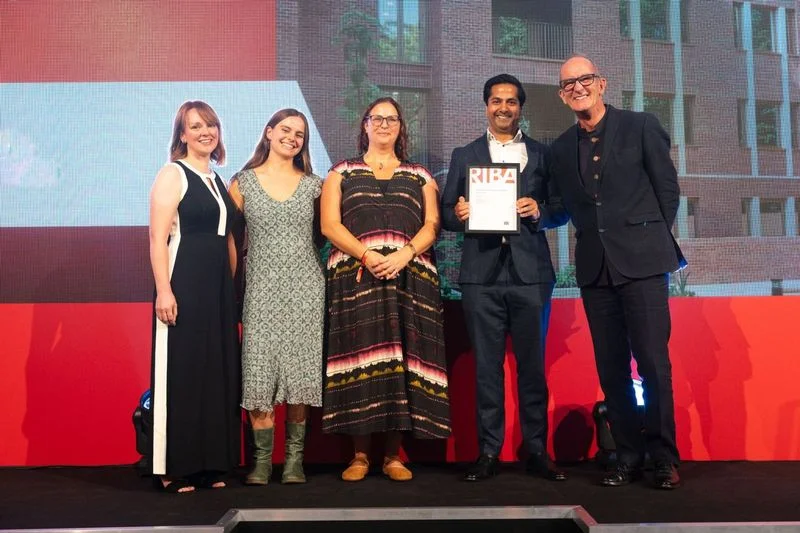 Five people on stage holding a RIBA award certificate, with a photograph of Rowan Court on the screen behind them.